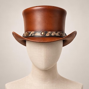 Puede incluir: Un sombrero de copa de color marrón medio liso con una corona estructurada y un ala curva. Una banda decorativa rodea la base de la corona, mostrando una textura con patrón de piel de serpiente acentuada con tachuelas metálicas circulares de color latón.
