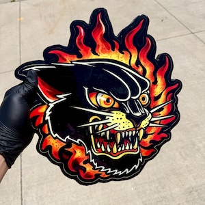May include: A die-cut sticker featuring a stylized black panther head with yellow eyes and bared teeth, surrounded by vibrant orange and red flames. The design uses bold black outlines and a glossy finish to create a high-contrast graphic illustration.