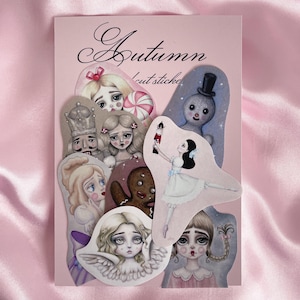 Puede incluir: Un conjunto de pegatinas troqueladas con ilustraciones de personajes, como una bailarina, un ángel y un hombre de jengibre, sobre un fondo rosa. La palabra "Autumn" está escrita en cursiva.