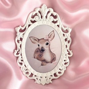 May include: An oval-shaped wall art piece featuring a painting of a fawn with a small pink bow on its ear. The artwork is encased in an ornate, white, scrollwork frame with intricate detailing along the edges and a pointed top and bottom.