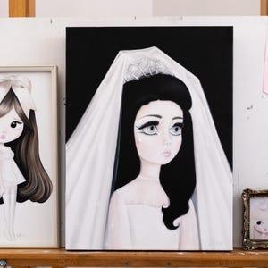 May include: Two framed art prints and one canvas painting. The left print shows a long-haired figure with a bow. The centre canvas depicts a figure in a white veil and tiara against a black background. The small right frame shows a similar figure.