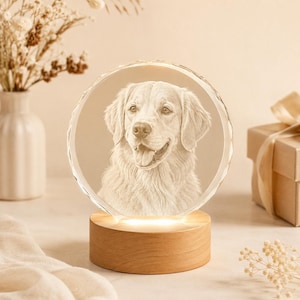 May include: A circular glass plaque featuring a laser-etched portrait of a golden retriever dog. The plaque is mounted on a cylindrical light-coloured wooden base that illuminates the glass from below, highlighting the detailed canine facial features.