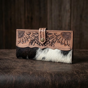 May include: A rectangular leather wallet featuring a top panel of tan leather with embossed sunflower patterns and a bottom panel of black and white cowhide fur. A central leather strap with a brass-colored metal snap button secures the wallet.