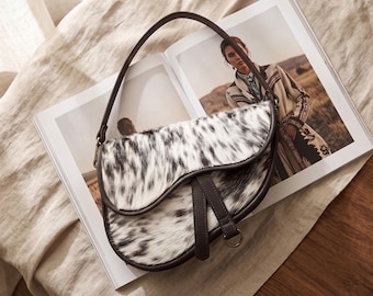 May include: A saddle-shaped shoulder bag featuring a white and black cowhide-style fur texture. The bag has a dark brown leather trim, a single matching leather shoulder strap, and a central leather strap detail with a metal D-ring attachment.