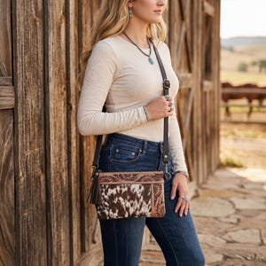May include: A crossbody bag featuring a brown and white cowhide panel, a dark brown leather section with an embossed floral pattern, a leather tassel, and an adjustable dark brown leather strap, worn with a long-sleeved cream shirt and jeans.