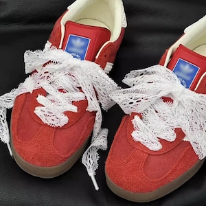 Könnte beinhalten: Ein Paar rote Wildleder-Sneaker mit weißen Spitzenbändern als Schnürsenkel. Die Sneaker haben eine flache Silhouette, eine Gummisohle und ein weißes Innenfutter. Ein blaues, rechteckiges Markenetikett ist auf der Zunge jedes Schuhs zu sehen.