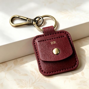 Könnte beinhalten: Ein quadratisches Schlüsselanhänger-Etui aus bordeauxfarbenem Leder mit einer Klappe und einem goldfarbenen Metalldruckknopf. Die Buchstaben BR sind auf die Klappe geprägt. Es enthält einen passenden goldfarbenen Schlüsselring und einen Karabiner.