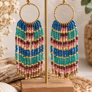 Könnte beinhalten: Ein Paar lange Fransenohrringe mit goldfarbenen, gehämmerten Metallringen und mehrfarbigen Glasperlen. Die Perlen sind in horizontalen Streifen aus Blau, Türkis, Rosa und Dunkelbraun angeordnet und bilden ein lebendiges Muster.