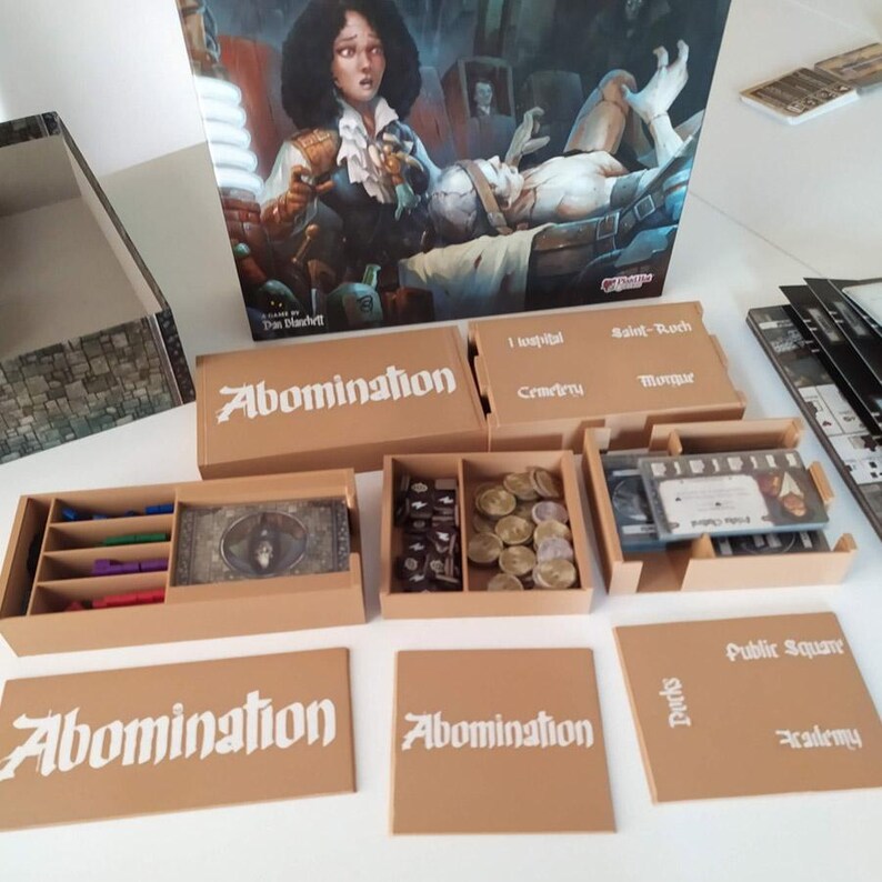 Puede incluir: Un juego de mesa titulado Abomination que incluye una tapa de caja grande ilustrada, varias bandejas de almacenamiento de pl&aacute;stico color canela y componentes de juego. Las bandejas contienen cubos negros peque&ntilde;os, monedas doradas y mazos de cartas. El texto incluye Abomination, Hospital, Saint-Roch, Cemetery, Morgue, Public Square, Docks y Academy.