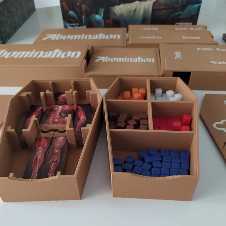 Puede incluir: Un organizador de juegos de mesa de pl&aacute;stico color canela con compartimentos en forma de ata&uacute;d. Una secci&oacute;n contiene una ilustraci&oacute;n de varias partes de una figura humanoide roja. Otra secci&oacute;n contiene cubos cuadrados en naranja, blanco, marr&oacute;n, rojo y azul.