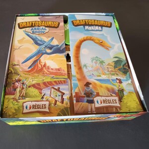 Pode incluir: Uma caixa de jogo de tabuleiro contendo dois manuais de regras para Draftosaurus Aerial Show e Draftosaurus Marina. O manual esquerdo mostra um pterossauro azul sobre um c&acirc;nion, enquanto o direito apresenta um dinossauro de pesco&ccedil;o longo.