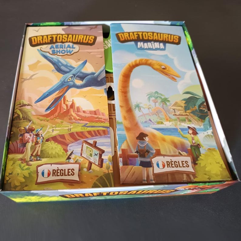 Pode incluir: Uma caixa de jogo de tabuleiro contendo dois manuais de regras para Draftosaurus Aerial Show e Draftosaurus Marina. O manual esquerdo mostra um pterossauro azul sobre um c&acirc;nion, enquanto o direito apresenta um dinossauro de pesco&ccedil;o longo.