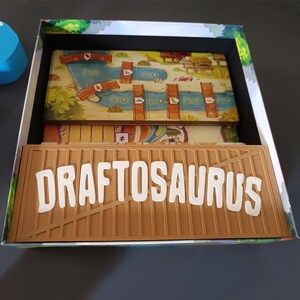 Pode incluir: Uma caixa de jogo de tabuleiro contendo uma bandeja de pl&aacute;stico bege com a palavra DRAFTOSAURUS em letras brancas em relevo. Abaixo da bandeja, um tabuleiro colorido apresenta um rio, pontes de madeira e &iacute;cones de dinossauros ilustrados.