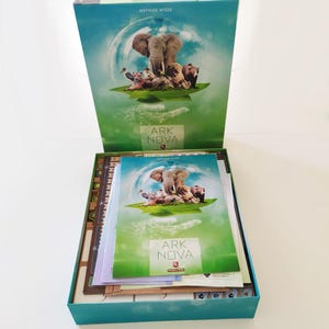 Op de afbeelding: De Ark Nova bordspeldoos is geopend en toont een spelregelboek met dezelfde afbeelding als het deksel. De cover toont een olifant, tijger, leeuw en beer op een groen blad tegen een blauwe lucht. De doos bevat spelborden en onderdelen.