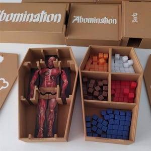 Puede incluir: Un juego de mesa en una caja color canela con forma de ata&uacute;d que contiene un rompecabezas de figura humana anat&oacute;mica roja y cuatro compartimentos con cubos peque&ntilde;os naranjas, blancos, marr&oacute;n oscuro, rojos y azules. La tapa muestra el texto Abomination.