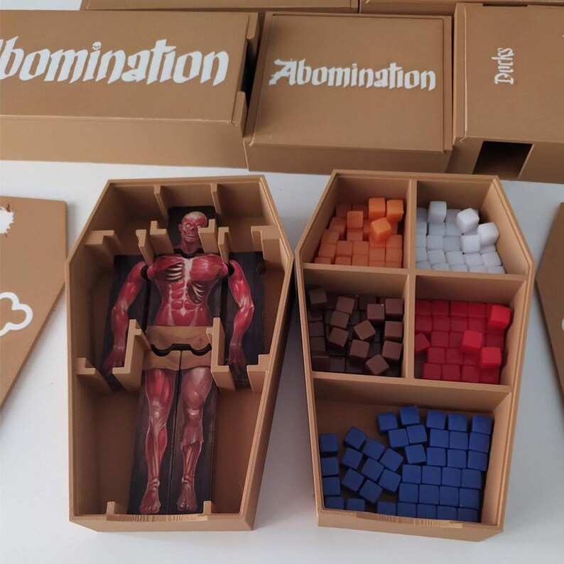 Puede incluir: Un juego de mesa en una caja color canela con forma de ata&uacute;d que contiene un rompecabezas de figura humana anat&oacute;mica roja y cuatro compartimentos con cubos peque&ntilde;os naranjas, blancos, marr&oacute;n oscuro, rojos y azules. La tapa muestra el texto Abomination.