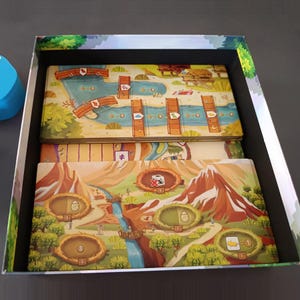 Pode incluir: Uma caixa de jogo de tabuleiro contendo dois tabuleiros retangulares com ilustra&ccedil;&otilde;es coloridas de paisagens, rios, montanhas e ninhos. Os tabuleiros apresentam caminhos numerados, &iacute;cones de pontes de madeira e v&aacute;rios s&iacute;mbolos gr&aacute;ficos de dinossauros.