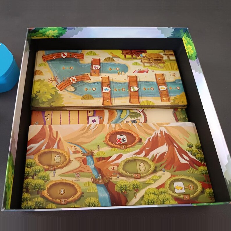 Pode incluir: Uma caixa de jogo de tabuleiro contendo dois tabuleiros retangulares com ilustra&ccedil;&otilde;es coloridas de paisagens, rios, montanhas e ninhos. Os tabuleiros apresentam caminhos numerados, &iacute;cones de pontes de madeira e v&aacute;rios s&iacute;mbolos gr&aacute;ficos de dinossauros.