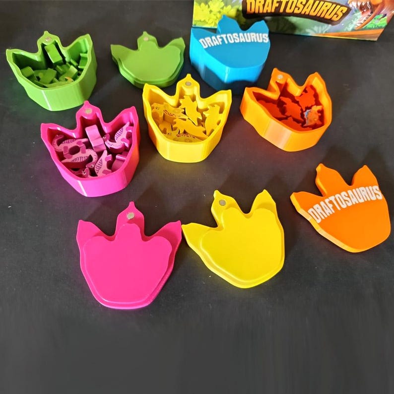 Pode incluir: Um conjunto de componentes do jogo de tabuleiro DRAFTOSAURUS com fichas em forma de dinossauro em v&aacute;rias cores. O set inclui v&aacute;rios recipientes em forma de pegada em rosa brilhante, amarelo, laranja, verde e azul, alguns com tampas e figuras.