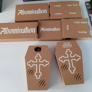 Puede incluir: Un conjunto de componentes de juego de color tostado que incluye cajas rectangulares etiquetadas como Abomination, Hospital, Saint-Roch, Cemetery, Morgue, Docks, Public Square y Academy. Dos cajas con forma de ata&uacute;d tienen cruces blancas.