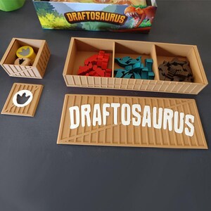 Pode incluir: Um organizador de jogo de tabuleiro em pl&aacute;stico castanho-claro para Draftosaurus com um tabuleiro retangular contendo fichas de dinossauro vermelhas, azul-petr&oacute;leo e castanhas. Um recipiente quadrado menor cont&eacute;m fichas de madeira amarelas e castanhas.