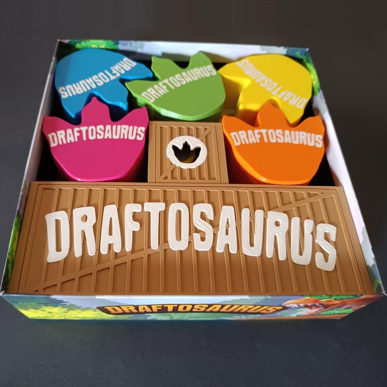 Pode incluir: Uma caixa de jogo de tabuleiro contendo fichas coloridas em forma de dinossauro nas cores azul, verde, amarelo, rosa e laranja, cada uma rotulada como DRAFTOSAURUS. Um divisor central marrom estilo engradado apresenta o texto DRAFTOSAURUS em letras brancas grandes.