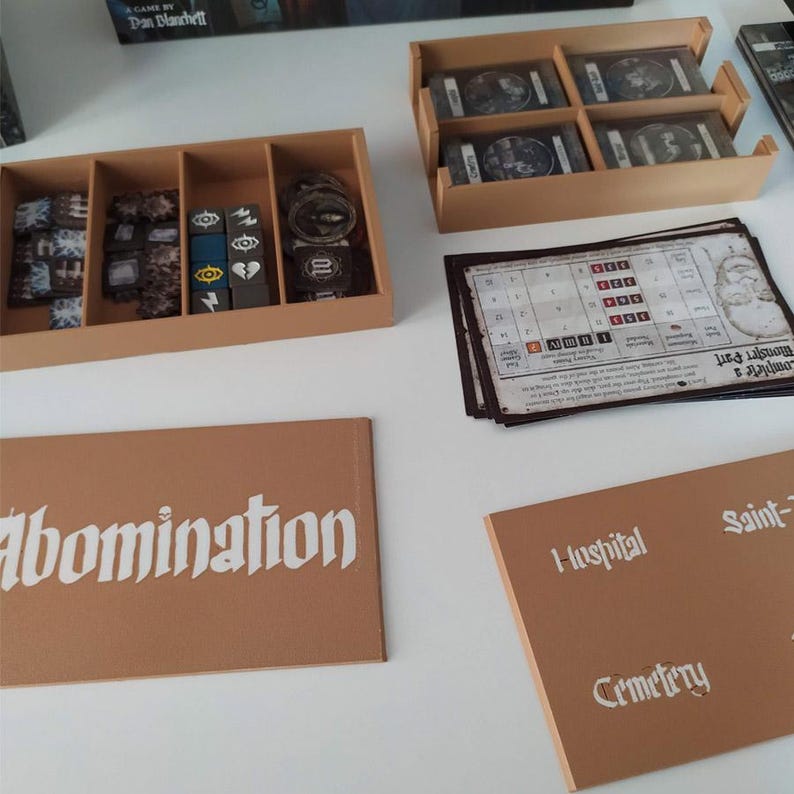 Puede incluir: Un conjunto organizador de juegos de mesa de pl&aacute;stico color canela que incluye una bandeja rectangular con cuatro compartimentos para fichas y dados, una bandeja cuadrada con cuatro ranuras para cartas y dos tapas planas con el texto Abomination, Hospital y Cemetery.