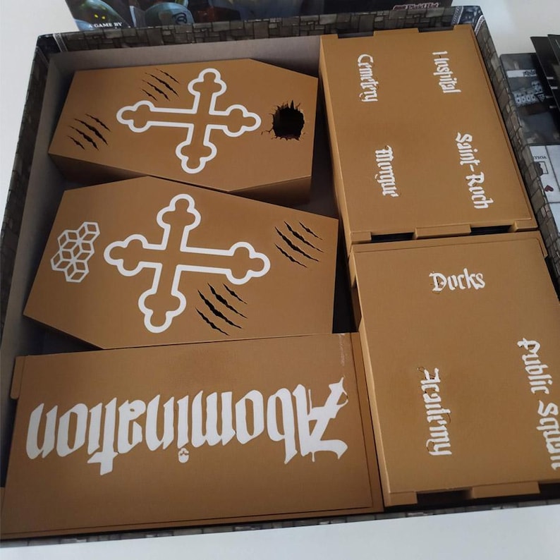 Puede incluir: El interior de una caja de juego de mesa con componentes de cart&oacute;n color canela. Dos piezas con forma de ata&uacute;d presentan gr&aacute;ficos de cruces blancas y marcas de garras. Las fichas rectangulares tienen impresos textos como Abomination, Cemetery, Hospital, Saint-Roch, Morgue, Docks, Academy y Public Square.