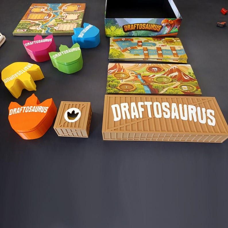 Pode incluir: Um conjunto de jogo de tabuleiro Draftosaurus com uma caixa, um tabuleiro, v&aacute;rios tabuleiros de jogador e recipientes coloridos em forma de dinossauro. Cada recipiente tem o texto Draftosaurus em branco, e a grande caixa retangular apresenta o mesmo texto em branco sobre um design de caixa bege.