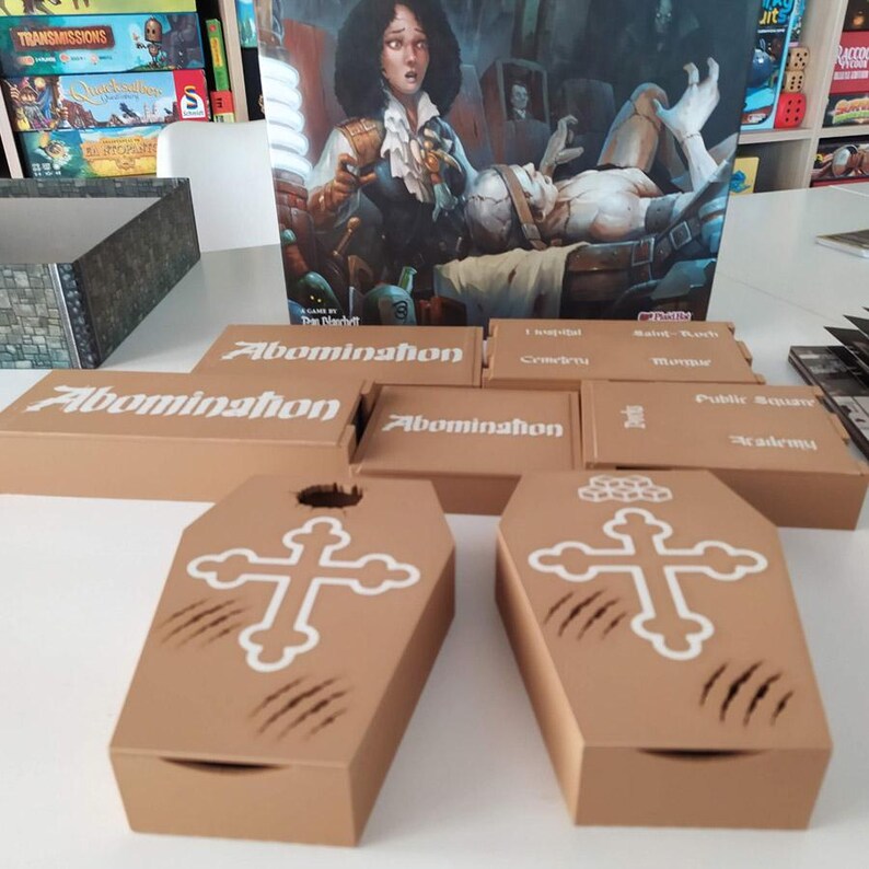 Puede incluir: Un juego de mesa que incluye una caja grande con una ilustraci&oacute;n g&oacute;tica, varias cajas de almacenamiento rectangulares de color canela etiquetadas como Abomination y dos cajas con forma de ata&uacute;d con cruces blancas y marcas de garras.