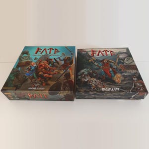 May include: Two rectangular board game boxes titled FATE. The left box is labelled Defenders of Grinheim by Jonathan Fryxelius, featuring fantasy characters in battle. The right box is labelled Monster Box, depicting a warrior fighting creatures.