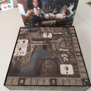 Puede incluir: Una caja de juego de mesa con una cubierta ilustrada oscura que muestra a una persona con bata de laboratorio y una figura p&aacute;lida sobre una mesa. El tablero interior muestra un mapa detallado de una ciudad con texturas de piedra y casillas.