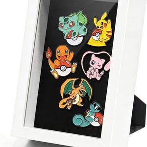 May include: A white shadow box frame containing six colorful enamel pins featuring various animated creatures. The pins include a green Bulbasaur, a yellow Pikachu, an orange Charmander, a pink Mew, an orange Charizard, and a blue Squirtle.