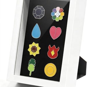 May include: A white rectangular display frame containing eight colorful enamel pins arranged on a black background. The pins feature various shapes including a dark gem, a multi-colored flower, a blue water drop, a pink heart, a yellow sunflower, a red flame, a green leaf, and a yellow circle.