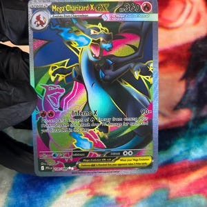 May include: A holographic Mega Charizard X EX trading card featuring a black dragon with blue flames, 360 HP, and the Inferno X attack. The card displays the text Mega Charizard X EX, 360 HP, Inferno X, and includes a small Charmander icon.