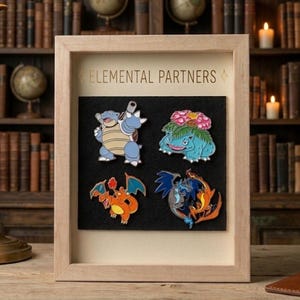 May include: A light-colored wooden shadow box frame containing four enamel pins on a black felt backing. The text "ELEMENTAL PARTNERS" is printed in gold at the top. The pins depict stylized blue, green, orange, and multi-colored creatures.