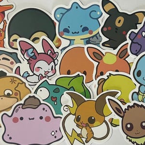 May include: A collection of die-cut vinyl stickers featuring various colorful cartoon creatures with large eyes and expressive faces, including orange, blue, pink, purple, and brown characters arranged in a dense, overlapping cluster.
