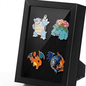 May include: A black rectangular display frame containing four enamel pins featuring colorful cartoon creatures. The pins depict a light blue turtle-like character, a green plant-based creature with a pink flower, an orange dragon, and a dark blue dragon.