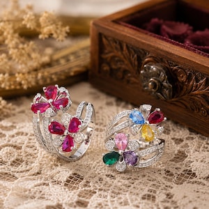 Könnte beinhalten: Zwei silberfarbene Ringe mit floralen Designs und birnenförmigen Edelsteinen. Ein Ring ist mit tiefrosa Steinen besetzt, während der andere eine bunte Anordnung aus blauen, gelben, rosafarbenen, violetten und grünen Steinen sowie kleinen klaren Kristallen zeigt.