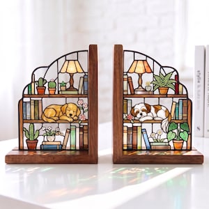 Puede incluir: Un par de sujetalibros de madera con inserciones de vidrieras. Cada pieza representa una estanter&iacute;a de varios niveles con un perro durmiendo, plantas en macetas, libros y una l&aacute;mpara. El lado izquierdo muestra un perro dorado y el derecho uno marr&oacute;n y blanco.