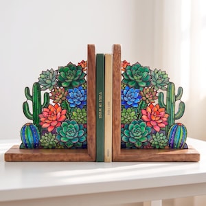 May include: A pair of wooden bookends featuring a colourful stained glass-style design of various succulents and cacti in shades of green, blue, orange, and red. Two books are positioned between the bookends, one with the text YUEJI IN MUSE.