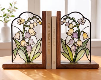 May include: A pair of bookends featuring stained glass panels with lily of the valley floral designs in shades of purple, yellow, and green, set within dark wood frames. Two books titled The Floral Archive and Garden Essays are held between them.