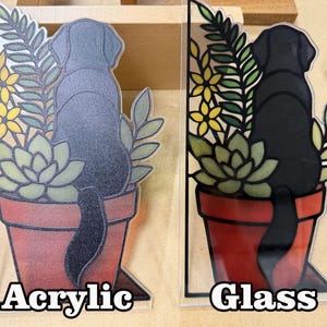 Puede incluir: Dos adornos decorativos con un perro negro sentado en una maceta de terracota con suculentas verdes y flores amarillas. Uno est&aacute; etiquetado como Acrylic con acabado esmerilado y el otro como Glass con un acabado liso y brillante.