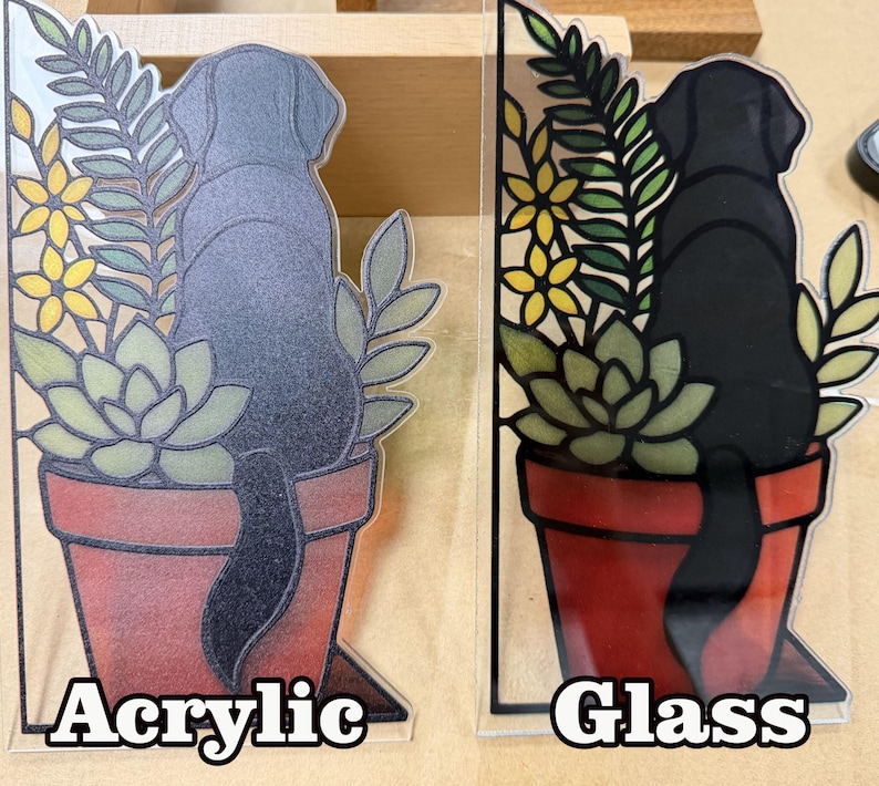 Puede incluir: Dos adornos decorativos con un perro negro sentado en una maceta de terracota con suculentas verdes y flores amarillas. Uno est&aacute; etiquetado como Acrylic con acabado esmerilado y el otro como Glass con un acabado liso y brillante.