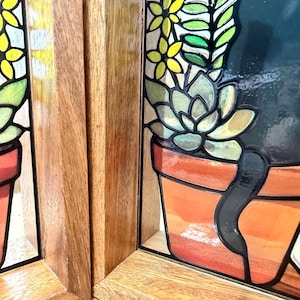 Puede incluir: Un panel de vidriera que muestra la silueta de un gato negro sentado junto a una planta suculenta en una maceta de terracota, con detalles florales amarillos y verdes. El arte en vidrio est&aacute; enmarcado en madera marr&oacute;n clara con veta vertical.