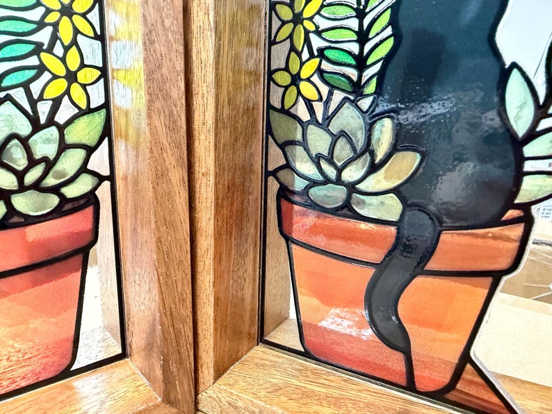 Puede incluir: Un panel de vidriera que muestra la silueta de un gato negro sentado junto a una planta suculenta en una maceta de terracota, con detalles florales amarillos y verdes. El arte en vidrio est&aacute; enmarcado en madera marr&oacute;n clara con veta vertical.