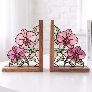 Könnte beinhalten: Ein Paar L-förmige Buchstützen mit einem Holzsockel und einer aufrechten Stütze. Jedes Stück zeigt ein Buntglasfenster mit rosa Orchideenblüten und grünen Blättern. Das Glas ist in einen dunklen Holzrahmen eingefasst und zeigt ein florales Motiv.