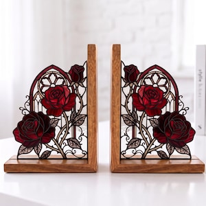 Puede incluir: Un par de sujetalibros en forma de L con base de madera e inserto de metal decorativo. El diseño metálico muestra rosas de color rojo intenso con hojas y tallos verdes, sobre un fondo de filigrana negra con arcos de estilo gótico.