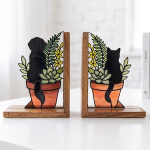 May include: A pair of L-shaped bookends featuring stained glass inserts set into wooden bases. One insert depicts a black dog sitting in a terracotta pot with green foliage and yellow flowers, while the other shows a black cat in a similar pot.