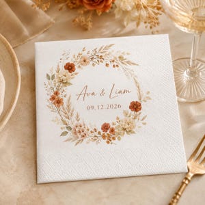 May include: A square white paper napkin featuring a printed floral wreath in shades of rust, cream, and beige. The center of the wreath displays the text "Ava & Liam" in a script font, followed by the date "09.12.2026" in a simple sans-serif font.